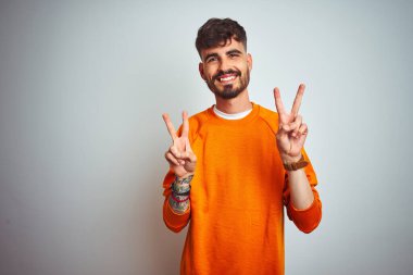 İzole edilmiş beyaz arka plan üzerinde turuncu kazak giyen genç adam kameraya bakıp zafer işareti yaparken gülümsüyor. İki numara..