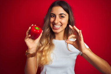 Kırmızı arka planda biber tutan genç ve güzel bir kadın. Parmaklarıyla imza atıyor. Mükemmel bir sembol.