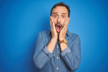 Yakışıklı, orta yaşlı, gri saçlı, mavi arka planı olan, korkmuş ve şok olmuş, şaşırmış ve yüzünde elleri olan yaşlı bir adam.