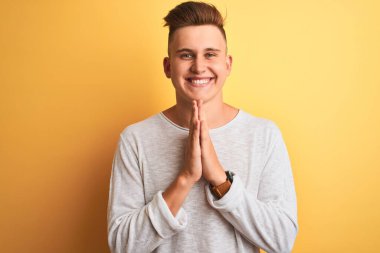 Beyaz tişört giyen yakışıklı genç adam izole edilmiş sarı arka planda duruyor ellerini birleştirip af diliyor kendinden emin bir şekilde gülümsüyor..