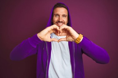 Spor tişörtü giyen genç fitness adamı ve mor izole edilmiş arka plan üzerine kapüşon takmış. Aşkla gülümseyen ve elleriyle kalp sembolü gösteren. Romantik konsept.
