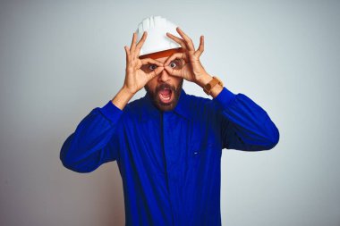 Üniformalı ve kasklı yakışıklı Hintli işçi izole edilmiş beyaz arka planda dürbün gibi hareket ediyor, gözleri parmaklarından bakıyor. Çılgın ifade.