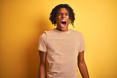Çizgili t-shirt giyen dreadlocks ile Afro adam izole sarı arka plan üzerinde duran kızgın ve deli çığlık sinirli ve öfkeli, öfke ile bağırarak. Öfke ve agresif kavram.