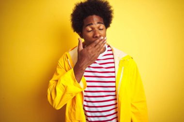 Siyah saçlı Afrikalı bir adam izole edilmiş sarı arka planda duran yağmurluk giymiş. Ağzı eliyle kapatmaktan sıkılmış. Huzursuz ve uykulu..