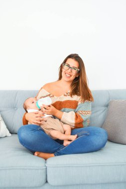 Genç, güzel bir kadın ve bebeği evdeki kanepede. Yeni doğan ve anne rahatlıyor ve dinleniyor. Beslenme şişesini kullanarak süt içiyorlar.