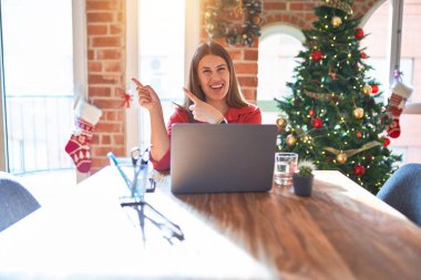 Güzel bir kadın evde dizüstü bilgisayarla oturmuş Noel ağacının etrafında gülümsüyor ve kameraya bakıyor iki eliyle ve parmaklarıyla işaret ediyor..