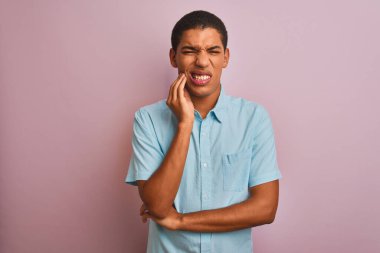 Mavi gömlekli genç yakışıklı Arap adam izole edilmiş pembe arka planda duruyor diş ağrısı ya da diş hastalığından dolayı acı dolu bir ifadeyle ağıza dokunuyor. Dişçi konsepti.