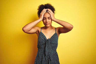 Afrika kökenli Amerikalı bir kadın, izole edilmiş sarı arka plan üzerine gündelik yeşil elbise giyip acı ve migren yüzünden çaresiz ve stresli bir baş ağrısı çekiyor. Ellerini başının üstüne koy..