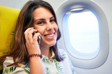 Havaalanında oturan genç yolcu kadın. Pencereden gökyüzü manzaralı. Akıllı telefon kullanarak arıyor.