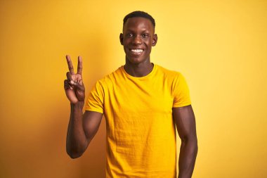 Sıradan tişörtler giyen genç Afro-Amerikan bir adam izole edilmiş sarı arka planda dikiliyor. Kameraya gülümsüyor ve parmaklarıyla zafer işareti yapıyor. İki numara..