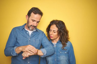 Güzel orta yaşlı çift, izole edilmiş sarı arka planda duruyorlar. Saati kontrol ediyorlar, rahat ve kendinden emin.