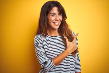 Çizgili tişört giyen genç ve güzel bir kadın. Sarı izole edilmiş arka planda neşeli duruyor. Yüzünde mutlu ve doğal bir ifade olan el ve parmak ile gülümseyen bir yüz var.