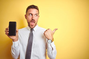 Genç yakışıklı iş adamı, sarı izole edilmiş arka planda akıllı telefon ekranı gösteriyor ve gülümseyen bir gülümsemeyle baş parmağıyla işaret ediyor.