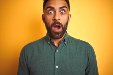 Yeşil tişörtlü genç Hintli adam izole edilmiş sarı arka planda duruyor. Şaşırmış ve korkmuş bir ifade, korku ve heyecanlı bir yüz ifadesi var..