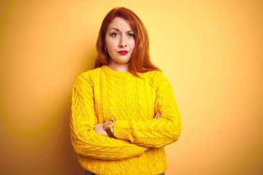 Kışlık süveter giyen güzel kızıl saçlı kadın izole edilmiş sarı arka plan üzerinde duruyor şüpheci ve gergin, yüzünde çapraz kollarla onaylamayan ifade. Negatif kişi.