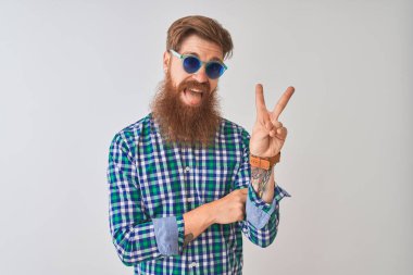 Genç, kızıl saçlı, günlük gömlek ve güneş gözlüğü takan, izole edilmiş beyaz arka planda gülümseyen ve kameraya gülümseyen, zafer işareti yapan bir adam. İki numara..