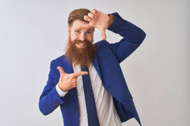 İzole edilmiş beyaz arka planda duran takım elbiseli genç kızıl saçlı işadamı gülümsüyor ve mutlu yüzlü elleri ve parmakları olan bir çerçeve yapıyor. Yaratıcılık ve fotoğrafçılık kavramı.