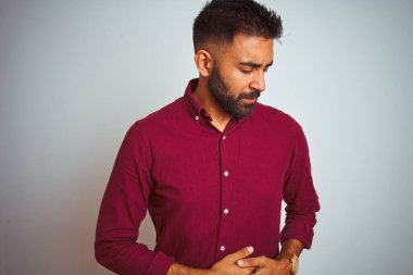 Kırmızı şık gömlek giyen genç Hintli adam midesi üzerinde gri arka planda duruyor çünkü hazımsızlık, acı verici bir hastalık kendini iyi hissetmiyor. Ache kavramı.