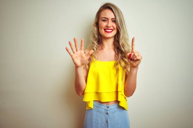 Sarı t-shirt giyen genç bir kadın beyaz izole edilmiş arka plan üzerinde duruyor ve altı numaralı parmaklarıyla işaret ederken kendinden emin ve mutlu gülümsüyor..