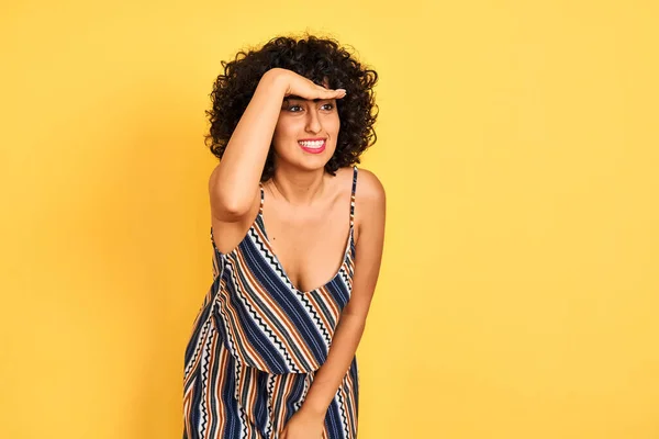 Arab woman with curly hair wearing striped colorful dress over isolated yellow background very happy and smiling looking far away with hand over head. Searching concept.