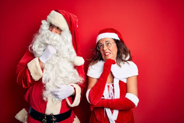Middle age couple wearing Santa costume and glasses over isolated red background Thinking worried about a question, concerned and nervous with hand on chin