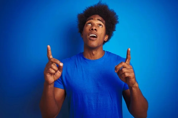 アフリカ系アメリカ人の男とアフロの髪を着てTシャツの上に立って隔離された青の背景に驚きと驚きを見上げ指と腕を上げて — ストック写真