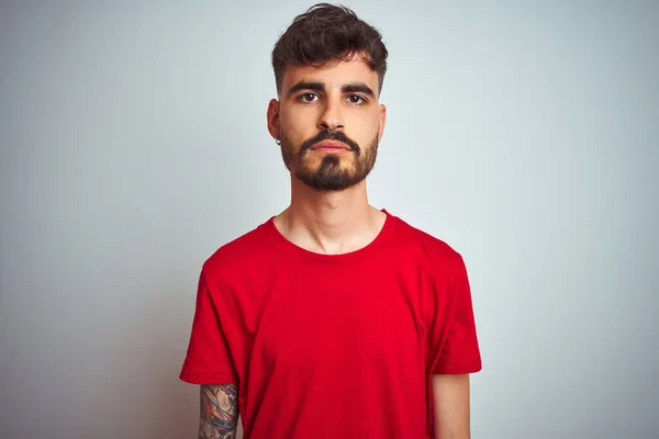 Young man with tattoo wearing red t-shirt standing over isolated white background Relaxed with serious expression on face. Simple and natural looking at the camera.