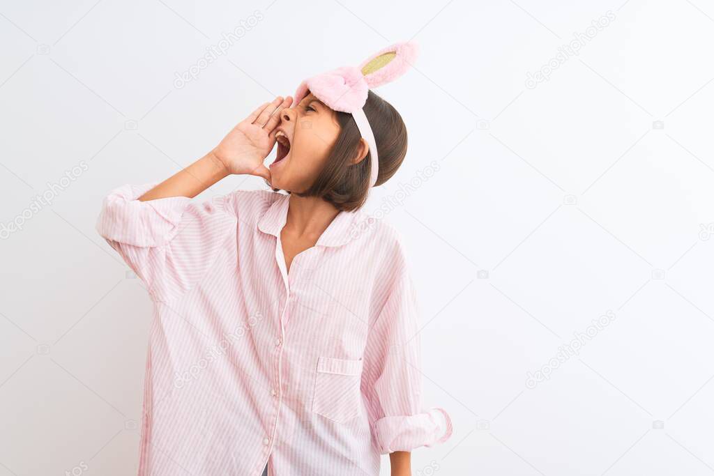 Beautiful child girl wearing sleep mask and pajama standing over isolated white background shouting and screaming loud to side with hand on mouth. Communication concept.