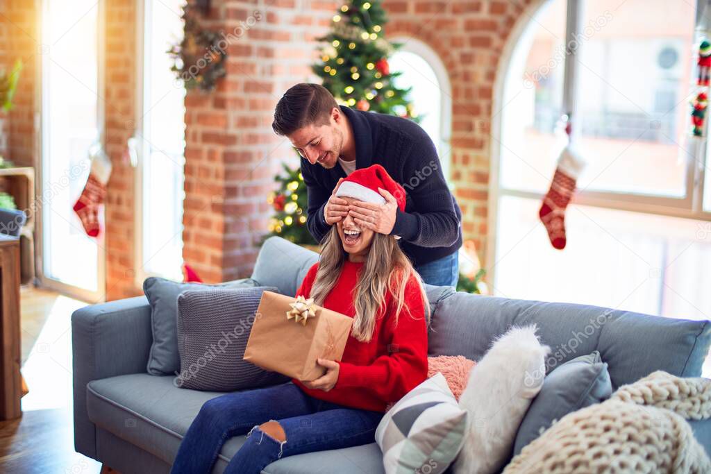 Young beautiful couple smiling happy and confident. Man covering eyes of woman surpise her with gift around christmas tree at home