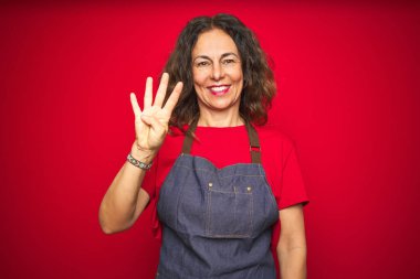 Kırmızı izole edilmiş arka planda önlük giyen orta yaşlı bir kadın dört numarayı işaret ederken kendinden emin ve mutlu bir şekilde gülümsüyor..