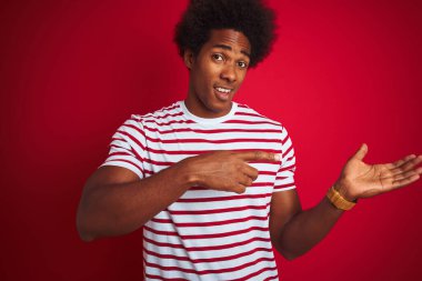 Afro saçlı, izole edilmiş kırmızı arka plan üzerinde çizgili t-shirt giyen genç Afro-Amerikan erkeği hayrete düşmüş ve kameraya gülümserken eliyle ve parmağıyla işaret ediyordu..