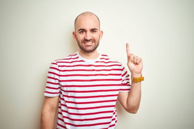 Sakallı, beyaz izole edilmiş arka plan üzerine sıradan kırmızı tişörtlü genç ve kel bir adam bir numarayı işaret ederken kendinden emin ve mutlu bir şekilde gülümsüyor..