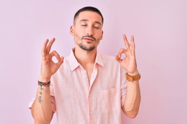 Pembe gömlekli genç adam izole edilmiş arka planda duruyor rahatla ve gözleri kapalı gülümse parmaklarıyla meditasyon hareketi yapıyor. Yoga konsepti.