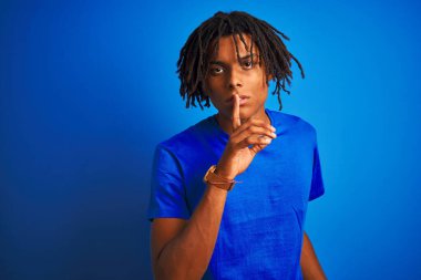 Rastalı tişörtlü Afro-Amerikan bir adam izole edilmiş mavi arka planda dikiliyor ve dudakları parmaklarıyla sessiz olmak istiyor. Sessizlik ve gizli konsept.