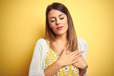 Sarı arka plan üzerinde duran ceketli güzel bir kadın. Elleri göğsünde, gözleri kapalı ve yüzünde minnet dolu bir jest ile gülümsüyor. Sağlık kavramı.