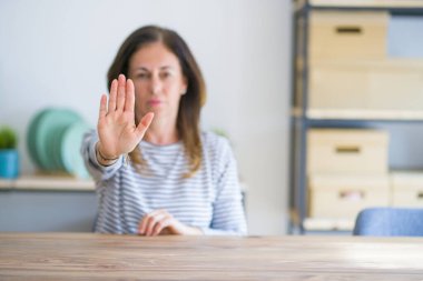 Orta yaş yaşlı kadın evde masada oturan avuç içi ile şarkı yapıyor. Yüzünde olumsuz ve ciddi bir jest olan uyarı ifadesi.