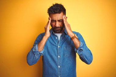 İzole edilmiş sarı arka plan üzerinde duran kot gömlekli genç Hintli adam. Stres yüzünden başının üstünde ağrı var. Migren ağrısı..