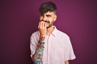 Yalıtılmış mor arka plan üzerinde duran pembe tişörtlü genç adam elleri ağız yiyen tırnaklarla gergin ve stresli görünüyor. Endişe sorunu.