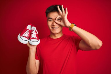 Asyalı genç bir Çinli spor ayakkabısı tutuyor. Kırmızı arka planda duruyor. Gülümsüyor, gülümsüyor.