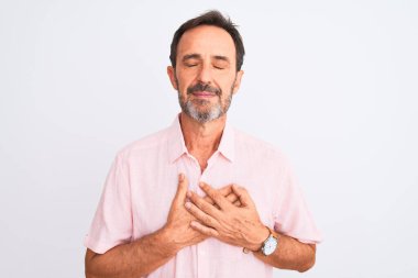 Orta yaşlı, pembe tişörtlü yakışıklı bir adam izole edilmiş beyaz arka plan üzerinde duruyor elleri göğsünde, gözleri kapalı ve yüzünde minnet dolu bir jest ile gülümsüyor. Sağlık kavramı.