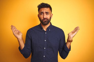 Genç ve yakışıklı Hintli işadamı, izole edilmiş sarı arka plan üzerinde gömlek giyiyor. Kolları ve elleri havada, kafası karışmış bir ifade takınıyor. Şüphe kavramı.