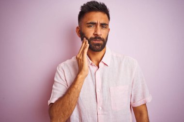 İzole edilmiş pembe arka plan üzerinde dikilen genç Hintli adam diş ağrısı ya da diş hastalığından dolayı acı dolu bir ifadeyle ağıza dokunuyor. Dişçi konsepti.
