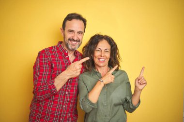 Izole sarı arka plan üzerinde Güzel orta yaş çift gülümseyerek ve yan iki el ve parmak ile işaret kamera bakıyor.