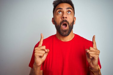 İzole edilmiş beyaz arka plan üzerinde kırmızı tişört giyen genç Hintli adam şaşırmış ve şaşırmış bir şekilde yukarı baktı ve parmaklarıyla işaret etti ve kollarını kaldırdı..