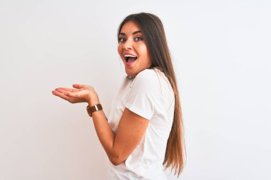 İzole edilmiş beyaz arka plan üzerinde duran sıradan bir tişört giyen genç ve güzel bir kadın avuç içleri açık bir şekilde fotokopi alanını gösteriyor, gülümseyerek mutlu bir şekilde gülümsüyor.