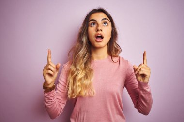 Pembe izole edilmiş arka planda süveter giyen güzel bir kadın şaşırmış ve şaşırmış bir şekilde yukarı bakıp parmaklarıyla işaret ediyor ve kollarını kaldırıyordu..