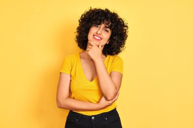 Sarı arka plan üzerinde duran kıvırcık saçlı genç Arap kadın çapraz kollarla gülümseyen ve çenesi üzerinde yükselen kameraya kendinden emin bir şekilde bakıyor. Olumlu düşünüyorum..