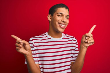 İzole edilmiş kırmızı arka plan üzerinde çizgili tişört giyen yakışıklı Arap adam parmaklarıyla farklı yönleri gösteren kendine güvenle gülümsüyor. Reklam için boşluk kopyala