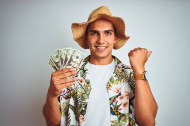 Tatilde elinde dolarlarla gezen genç bir adam izole edilmiş beyaz bir arkaplanı işaret ediyor ve gülümseyen bir yüzle baş parmağıyla onu gösteriyor.