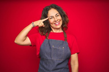 Kırmızı izole edilmiş arka planda önlük giyen orta yaşlı bir kadın tek parmağıyla gülümseyerek, harika bir fikir ya da düşünceyle, iyi bir hafızaya sahip.